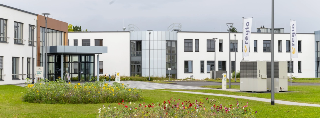 Modern white office building with glass entrance, landscaped garden, and outdoor equipment units.