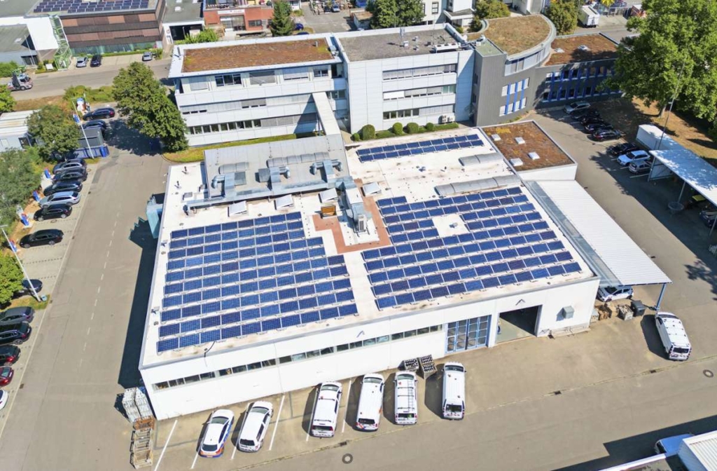 Aerial view of a large industrial building with numerous solar panels installed on its flat roof. The building is surrounded by parked cars and other structures with green roofs and trees nearby