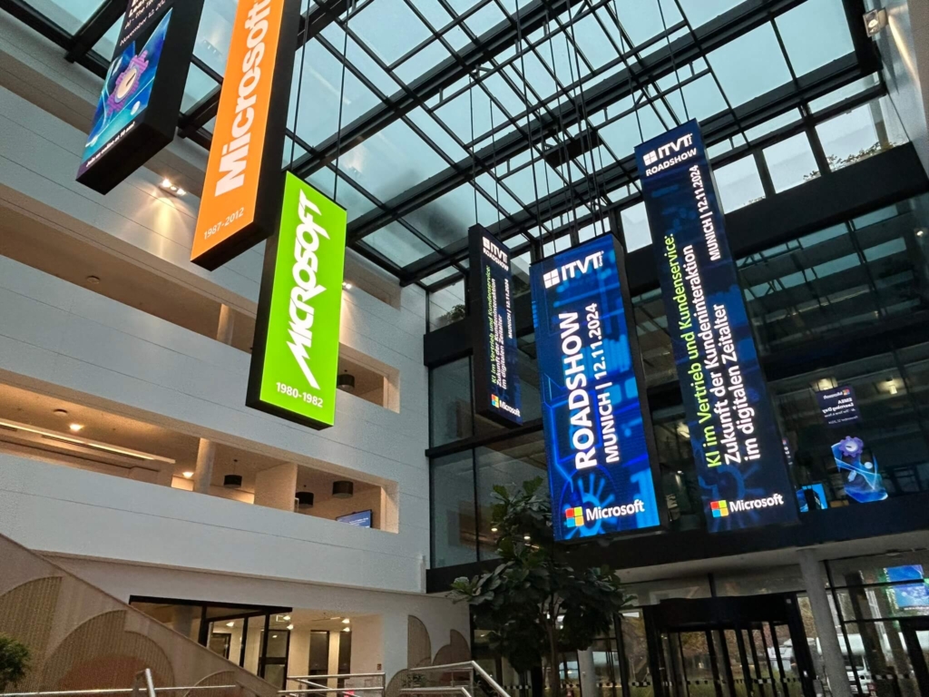 Large indoor atrium with hanging vertical banners displaying "Microsoft" logos and an ITVT roadshow announcement for Munich, dated 12.11.2024, promoting AI in sales and customer service.