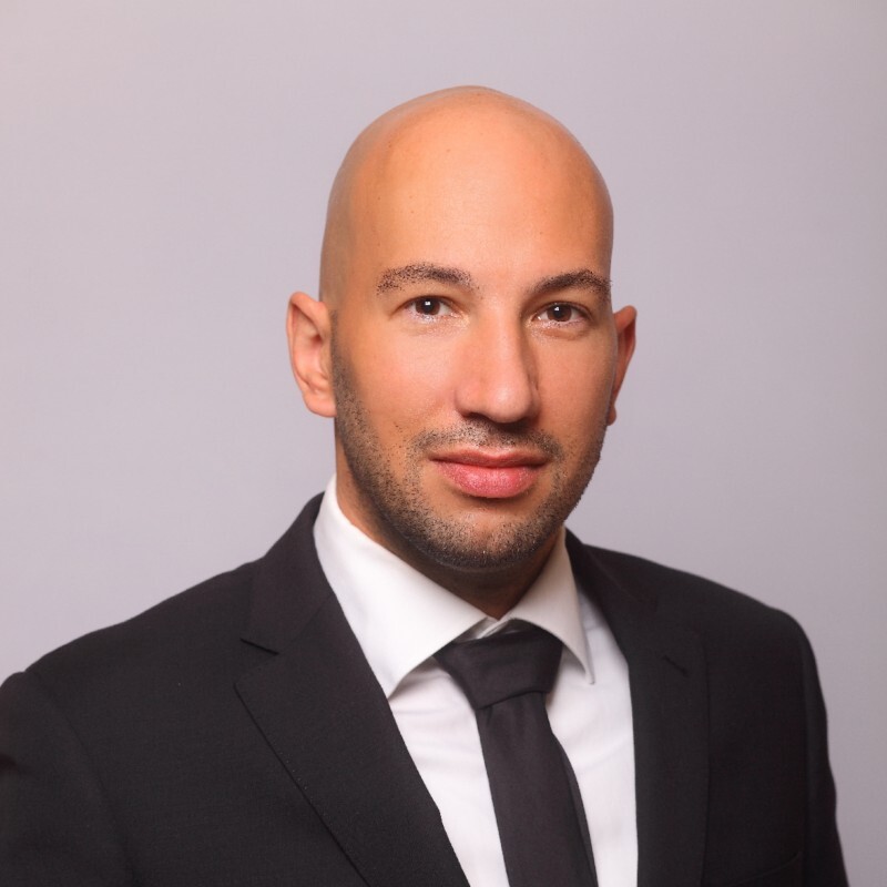 Ein glatzköpfiger Mann in Anzug und Krawatte, der selbstbewusst und mit neutraler Miene dasteht.