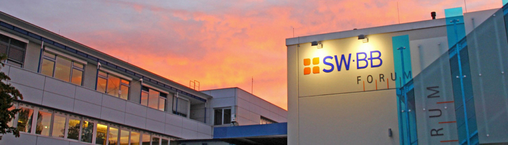 Modernes Gebäude mit beleuchtetem SWBB-Forum-Logo an der Fassade, unter einem farbenfrohen Sonnenuntergangshimmel mit orangefarbenen und rosafarbenen Tönen.