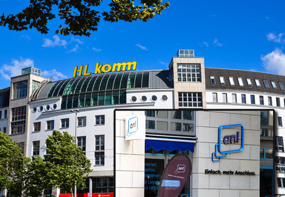 HL-Komm-Gebäude mit gewölbtem Glasdach und Beschilderung, mit zusätzlicher Ladenfront mit dem Branding „en! Einfach. mehr Anschluss.“ unter strahlend blauem Himmel.
