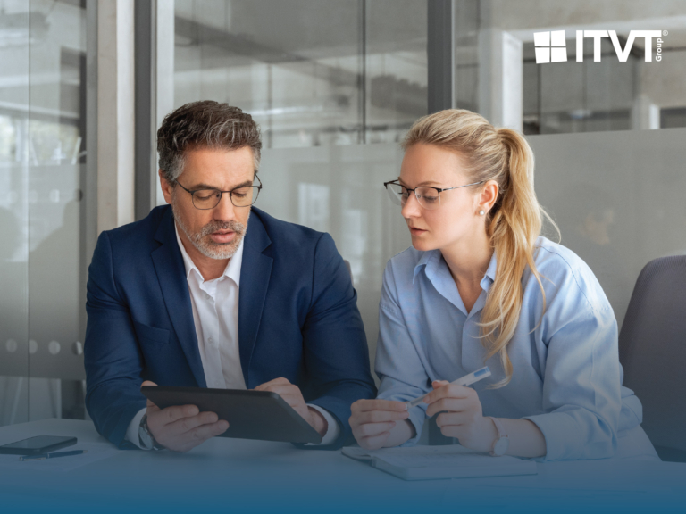 Zwei Geschäftsleute, ein Mann in einem blauen Anzug und eine Frau in einem hellblauen Hemd, beide mit Brille, überprüfen Informationen auf einem Tablet in einer Büroumgebung. Das Logo der ITVT Group ist in der oberen rechten Ecke zu sehen.